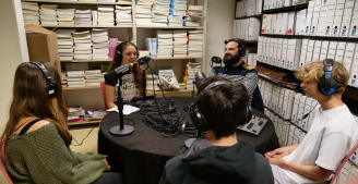 On voit cinq personnes assises autour d’une table ronde, avec des casques sur les oreilles et des micros devant elles. Les lycéens sont en train d'échanger avec un professionnel pour le podcast dans une pièce remplie de livres et de boîtes d’archives.